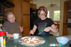Scout Son and Snarky Daughter dollop cheese onto the pizza